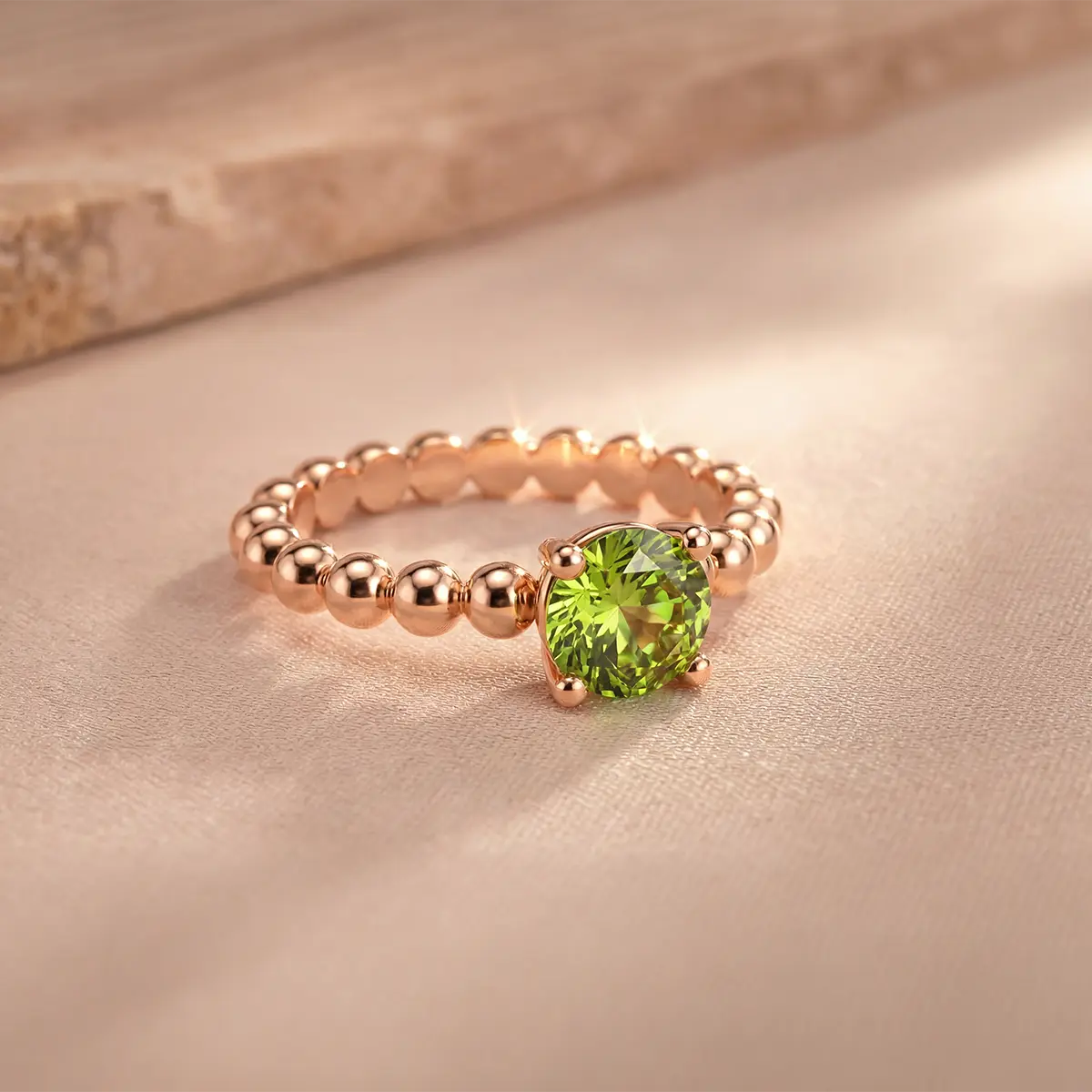 Bague solitaire en or rose 18k sertie d'un péridot rond vibrant sur un anneau perlé signature, posée sur un fond texturé doux, illustrant l'élégance de la collection Bagues Péridot Bajoia