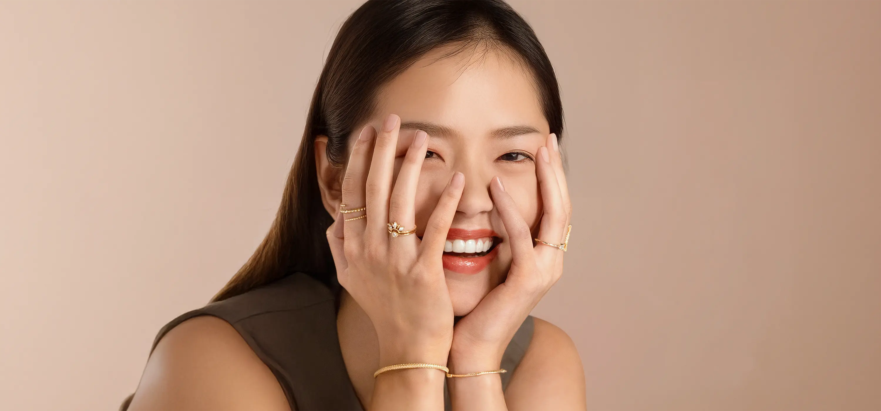 Femme souriante portant une accumulation de bagues fantaisie dorées et bracelets fins Bajoia, mains posées sur le visage mettant en valeur l’éclat des bijoux.