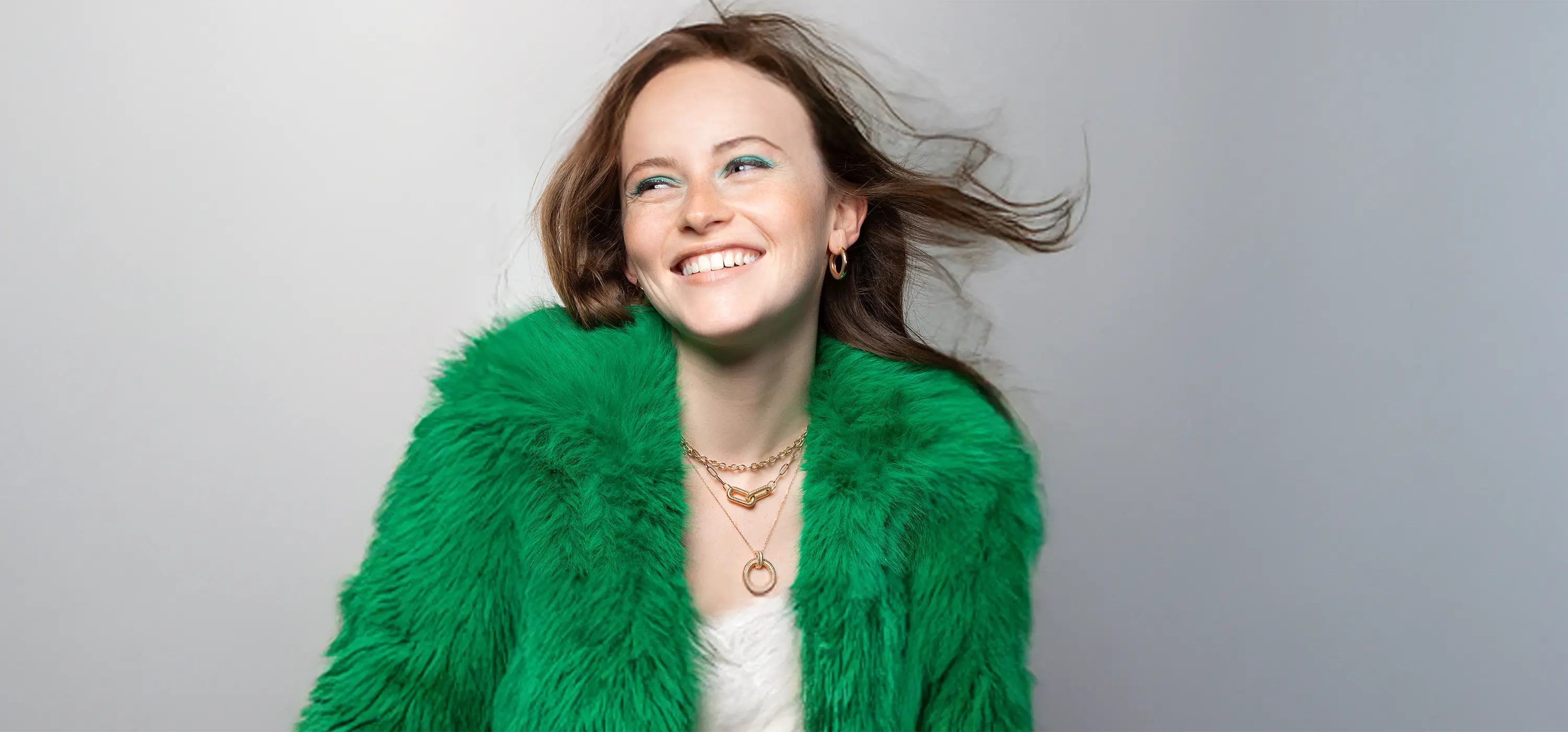 Femme souriante portant un manteau vert et une accumulation de colliers dorés Bajoia : ras-de-cou chaîne et pendentif anneau texturé.